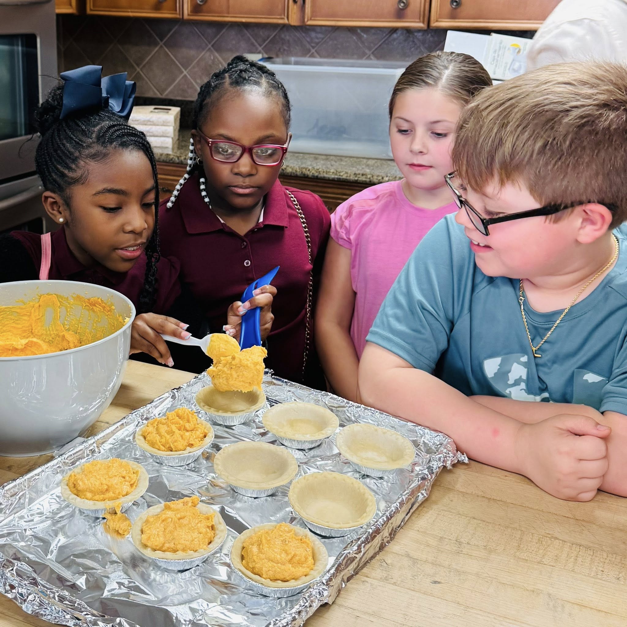 A boy and two girls watching another girl put pie filling into mini pie shells.